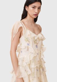 Blomstermønstret kjole i lys cremefarvet stof, med lagdelte flæser på ærmerne og bodice, fine sting og et blødt fald.