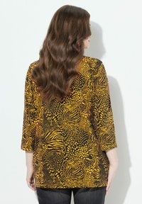 Mustan ja keltaisen kuvioitu paita, jossa on kolme neljäsosaa hihat, ja joka on koristeltu abstrakteilla pyörteillä ja pilkuilla. Paita on valmistettu sileästä kankaasta.