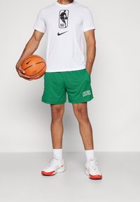Vit Nike t-shirt med NBA-logotyp, gröna mesh-shorts med texten "CELTICS", orange basketboll och vita/röda träningsskor med ankelsockar.