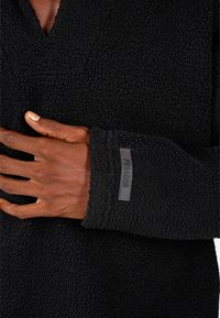 Čierny fleecový pulóver s V-výstrihom, textúrovanou tkaninou a prišitým štítkom, na ktorom je napísané "PECADOR" na manžete.