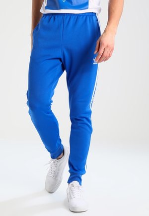 Mann trägt blaue Adidas-Trainingshose mit weißen Streifen und weiße Turnschuhe vor einem schlichten hellen Hintergrund.