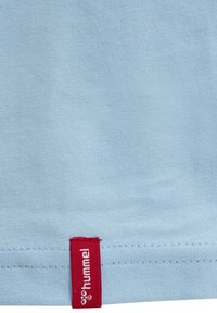 Lyseblåt stof med en rød påsyet mærkat med det hvide "hummel" brandlogo og tekst nær kanten.