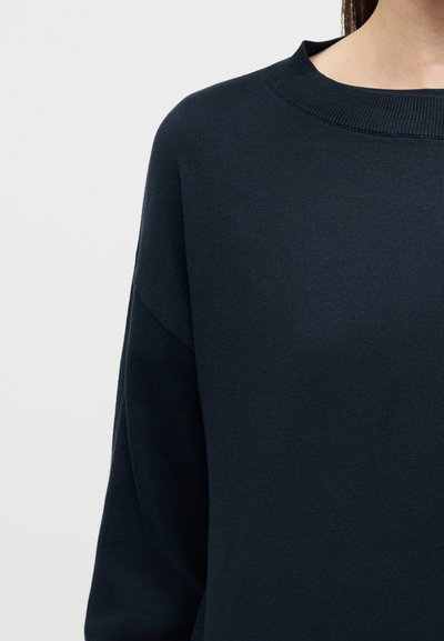 Γυναίκα που φοράει σκούρο μπλε πουλόβερ με στρογγυλή λαιμόκοψη, κομμένο στους ώμους και τον λαιμό, σε απλό ανοιχτό φόντο.