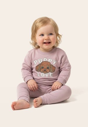 Uśmiechnięte małe dziecko o blond włosach siedzące boso, ubrane w różowy strój z twarzą szczeniaka i napisem „Puppy Love” na bluzie.