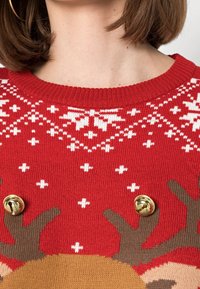 Pull rouge en tricot avec des motifs de flocons de neige blancs et un motif de renne. Des clochettes dorées accentuent le design. Texture de tricot circulaire sur toute la surface.