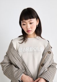 Beige pulóver "BARBOUR INTERNATIONAL" hímzett felirattal, egy barna és bézs kockás gombos kabát alatt viselve. Sima textúra.