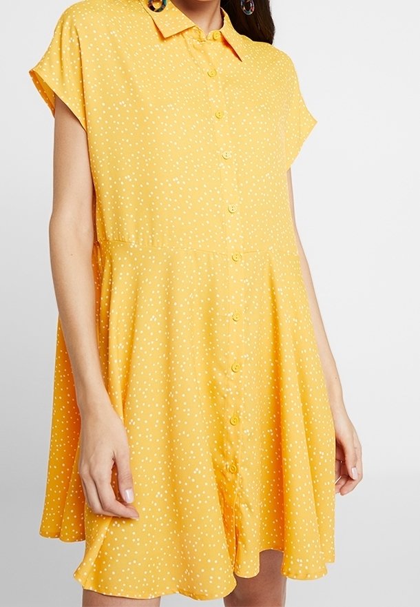 Femme portant une robe jaune à manches courtes avec des pois blancs, une fermeture à boutons sur le devant, et une jupe ample évasée.