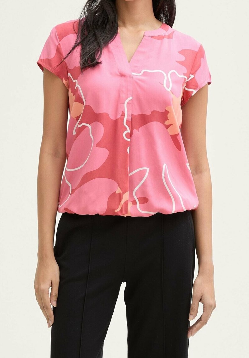 Femme portant un chemisier à manches courtes rose à motif floral et un pantalon noir, vue des épaules aux hanches sur un fond clair.