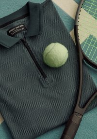 Polo à zip mi-long vert foncé texturé avec une balle de tennis posée dessus et une raquette de tennis noire aux cordes vert néon sur un tapis bleu et beige.