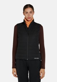 Kvinna som bär en svart vadderad väst över en brun långärmad topp och en orange halsduk, stående mot en enkel grå bakgrund.