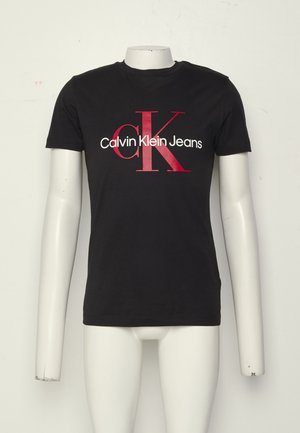 Sort T-shirt med korte ærmer med røde "cK" bogstaver og hvid "Calvin Klein Jeans" tekst foran, vist på hovedløs mannequin.