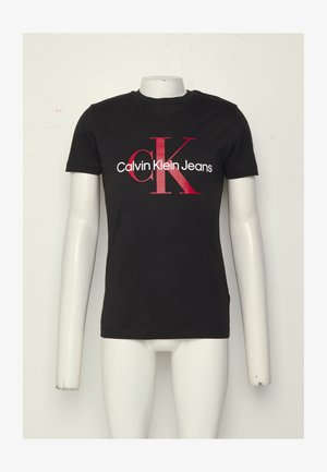 Černé tričko s krátkým rukávem s červenými písmeny "cK" a bílým nápisem "Calvin Klein Jeans" na přední straně, vystavené na figuríně bez hlavy.