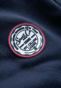 Rund hvid og sort stofmærke med rød kant, tekst "Sport and Co. 1985 NYC College League" på mørkeblåt stofbaggrund.