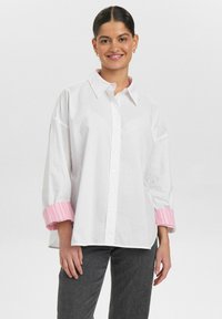 Camicia bianca con colletto grande e polsini a strisce rosa. Il tessuto appare liscio e la camicia ha un taglio morbido.