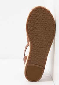 Semelle de sandale marron avec un motif répété du logo "MK" et une prise de talon texturée, inclinée contre une surface blanche.