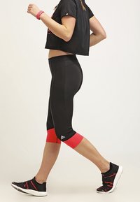 Kvinde der går i sorte cropped leggings med røde manchetter, sort cropped T-shirt, sorte og røde sneakers samt en pink fitness tracker på venstre håndled.