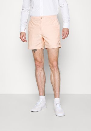 Uomo che indossa pantaloncini rosa chiaro, camicia bianca a maniche lunghe, calzini bianchi e scarpe da ginnastica bianche in piedi davanti a uno sfondo semplice.