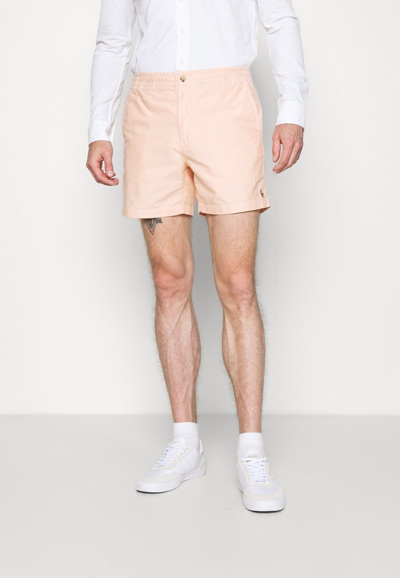 Homme portant un short rose clair, une chemise blanche à manches longues, des chaussettes blanches et des baskets blanches, debout devant un fond uni.