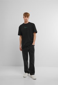 T-shirt nera in cotone a maniche corte con un logo verde sul petto, abbinata a pantaloni grigio scuro e sneakers bianche.