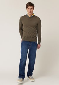 Maglione a collo rotondo verde oliva, indossato sopra una camicia a colletto blu e gialla. Abbinato a jeans blu e sneaker beige.
