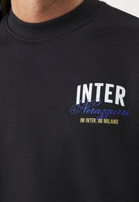 Gros plan d’un t-shirt noir à col rond avec le texte "INTER Nerazzurri" et "IM INTER, IM MILANO" en blanc, bleu et jaune sur la poitrine.