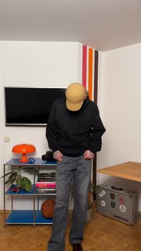 Άτομο με καπέλο καφέ ανοιχτό και μαύρο φούτερ κοιτάζει προς τα κάτω, στέκεται σε δωμάτιο με τηλεόραση, πορτοκαλί φωτιστικό, μπάλα του μπάσκετ, βιβλία και μια βαλίτσα με αυτοκόλλητα.