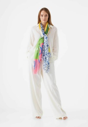 Chemise blanche texturée à manches longues portée sous une écharpe multicolore. L'écharpe présente des motifs géométriques rose, vert et bleu. Pantalon blanc à jambes larges.