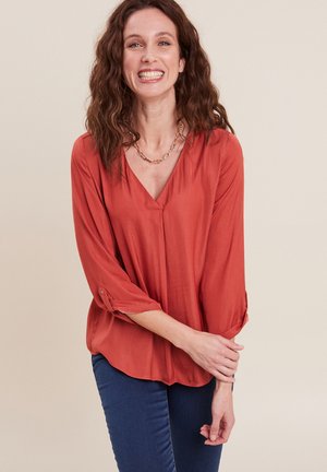 Blusa leggera di color ruggine con scollatura a V, maniche a tre quarti con risvolti e orlo curvo. Indossata con jeans blu.