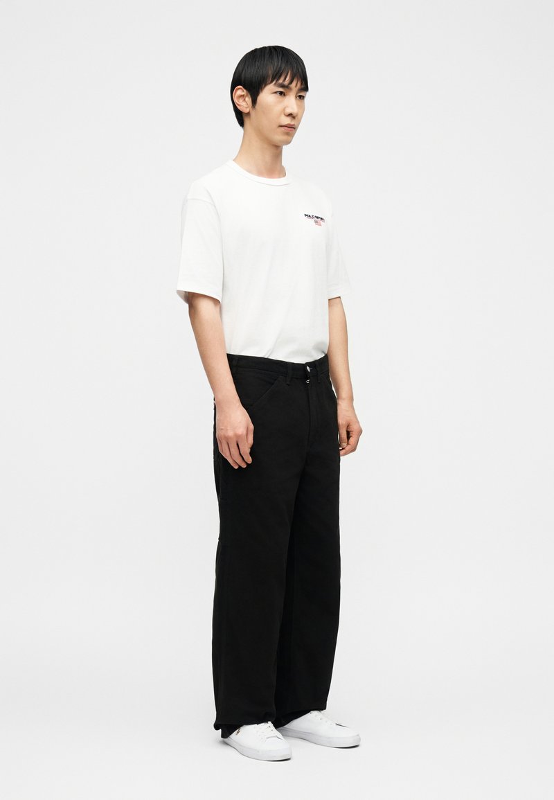 Homme debout portant un T-shirt blanc à motif, un pantalon noir ample et des baskets blanches, sur un fond blanc uni.