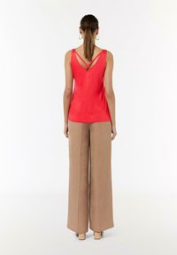 Czerwony bezrękawnik z dekoltem w kształcie litery V i krzyżującymi się paskami na plecach, w połączeniu z beżowymi spodniami o szerokich nogawkach, widok od tyłu.