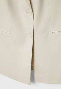Veste blanche cassée avec deux boutons et deux poches horizontales fendues à l'avant, vue rapprochée de la partie basse du torse.
