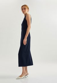 Marineblaues ärmelloses Kleid mit einer taillierten Oberseite und einem A-Linie-Rock, der eine glatte Textur aufweist. Kombiniert mit weißen Slingback-Pumps mit spitzem Zehenbereich.
