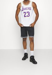 Weißes Basketballtrikot mit "Lakers"-Text und der Nummer "23" in Lila, kombiniert mit schwarzen Shorts mit weißen Streifen und schwarzen Turnschuhen.