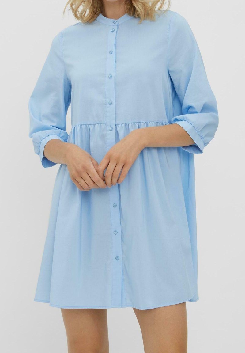 Femme portant une robe bleu clair à manches longues boutonnée, taille haute et jupe froncée, debout les mains jointes.