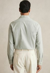 Cămașă lungă cu mâneci verzi deschis, cu o textură netedă, cu manșete butonate, croială dreaptă, purtată băgată în pantaloni de culoare crem.