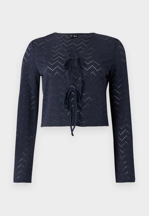 Tmavě modrá halenka s dlouhým rukávem s chevronovým vzorem s očky, výstřihem do V a dvěma uvázanými mašlemi uprostřed vpředu.