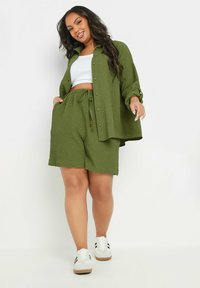 Set in tessuto verde a due pezzi composto da una camicia con bottoni e pantaloni corti morbidi, abbinato a sneakers bianche. Maniche corte e vita con coulisse.