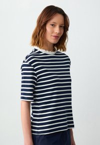 Jonge vrouw met schouderlang bruin haar, gekleed in een marineblauwe en witte horizontaal gestreepte shirt met een witte, geschulpte kraag.