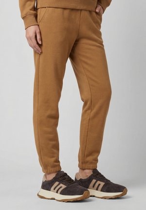 Dolna część ciała ubrana w brązowe spodnie dresowe i ciemnobrązowe trampki z beżowymi akcentami na jasnoszarym tle.