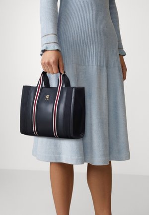 Černá kožená kabelka s bílými a červenými proužky, obdélníkového tvaru, s pevnými uchy a zlatým logem na přední straně.