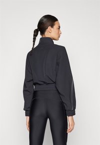 Black cropped jacket with a high collar and elastic waistband, featuring long sleeves and textured fabric. Worn with black leggings.