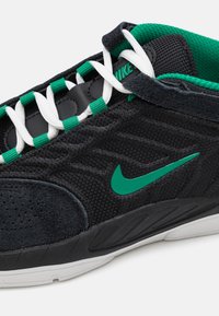 A Nike cipők fekete hálós felsőrésze zöld részletekkel, fehér fűzőkkel és texturált fekete velúrból készült orral rendelkeznek. Az outsole főként fehér színű.