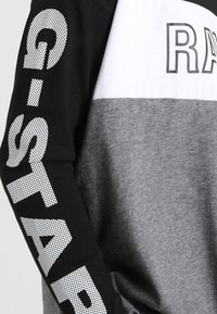 Långärmad skjorta med svarta ärmar och vita, grå färgblock, med vit prickig text "G-STAR" på ärmen och delvis "RA" på bröstet.