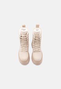 Μπεζ χειμερινές μπότες με χοντρές σόλες, κορδόνια μπροστά και λευκή γούνα στο εσωτερικό, εμφανίζονται από πάνω σε ανοιχτό φόντο.
