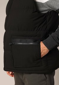 Zwarte gewatteerde vest met een ritszak aan de voorkant en zijnaaddetails. Het materiaal heeft een gladde textuur met subtiele stiksels.