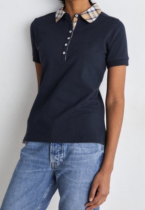 Persoon draagt een marineblauw polohemd met korte mouwen, een beige geruite kraag en lichtblauwe spijkerbroek, staand met één hand ontspannen langs het lichaam.