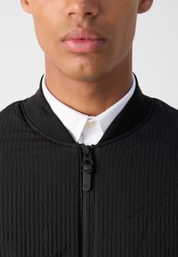 Svart quiltet jakke med høy krage, med glidelås foran og en hvit skjorte under. Teksturert stoff og minimalistisk design.