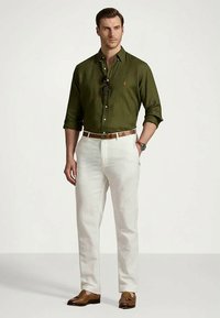 Homem vestindo uma camisa de botão verde-oliva, calças brancas, cinto castanho, sapatos de estilo loafers com franjas castanhas e óculos de sol pendurados na gola da camisa.