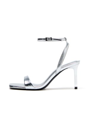 Srebrni kovinski sandal z visoko peto, zanko za gleženj in zaponko, odprti prsti in tanko peto na belem ozadju.