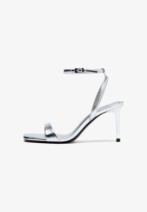 Srebrni kovinski sandal z visoko peto, zanko za gleženj in zaponko, odprti prsti in tanko peto na belem ozadju.